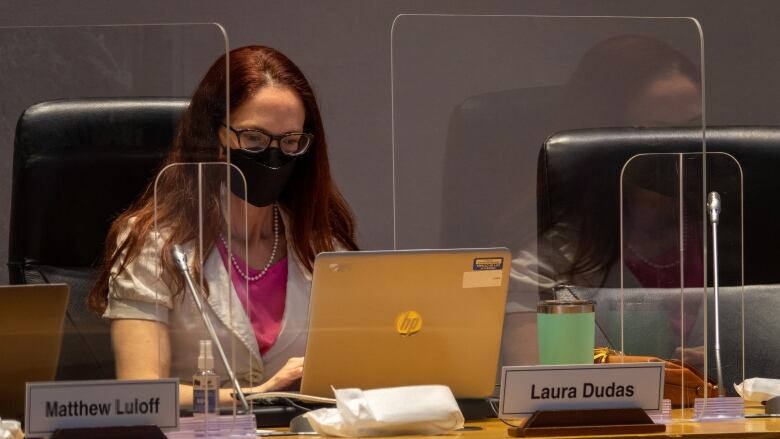 A woman in a mask looks down at her laptop while sitting at a desk behind plexiglass.