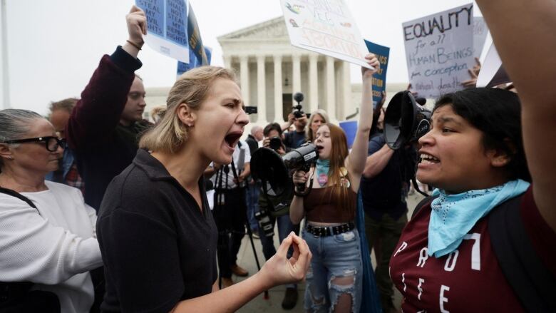 Two women stand face-to-face, shouting at each other, in the midst of a large crowd gathered outside a courthouse.