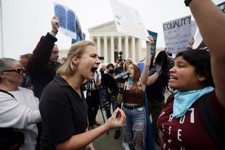 Two women stand face-to-face, shouting at each other, in the midst of a large crowd gathered outside a courthouse.