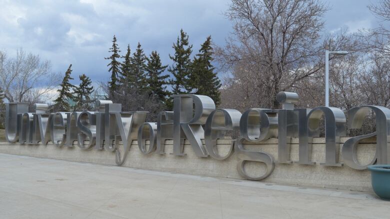 The words 'University of Regina' are made of silver, etched into a stone wall. Behind the sign are trees and a cloudy sky.