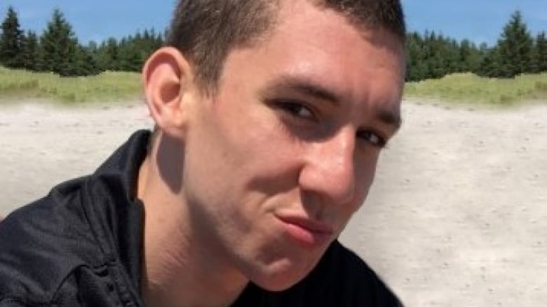 A man with a buzzcut poses for a photo while sitting down on a sandy beach.