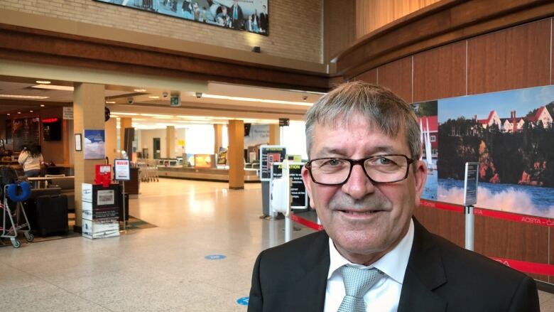 A man with glasses in a dark suit, white shirt and light blue tie stands in the airport waiting area.