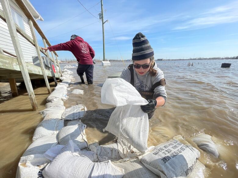 Two people wade in water as they place sandbags in a line around a house.