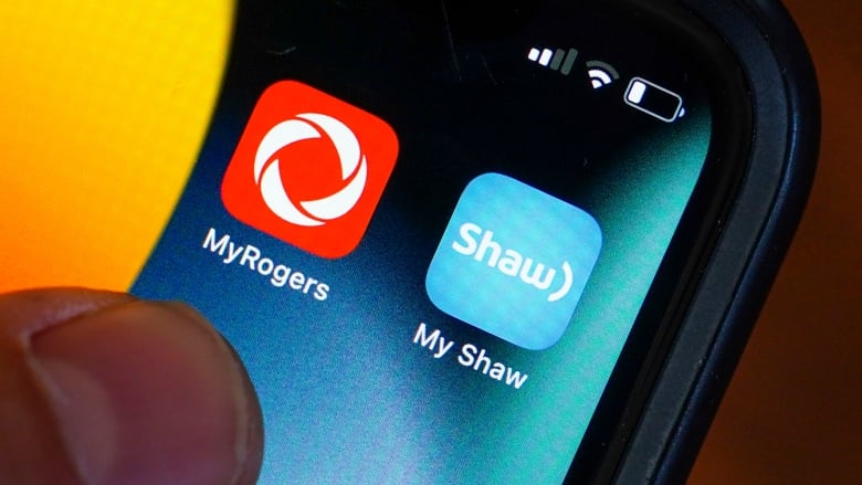 The logos of Rogers Communications and Shaw Communications appear on a cellphone screen. 