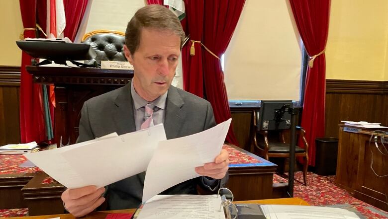 Man with short brown hair wearing grey suit jacket with tie sits at a desk in a city council room looking through papers.