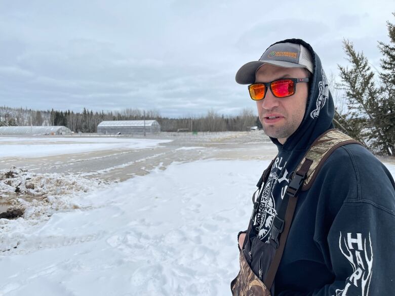 A man in sunglasses and a hoodie stands beside a snowy field with greenhouses visible in the background.