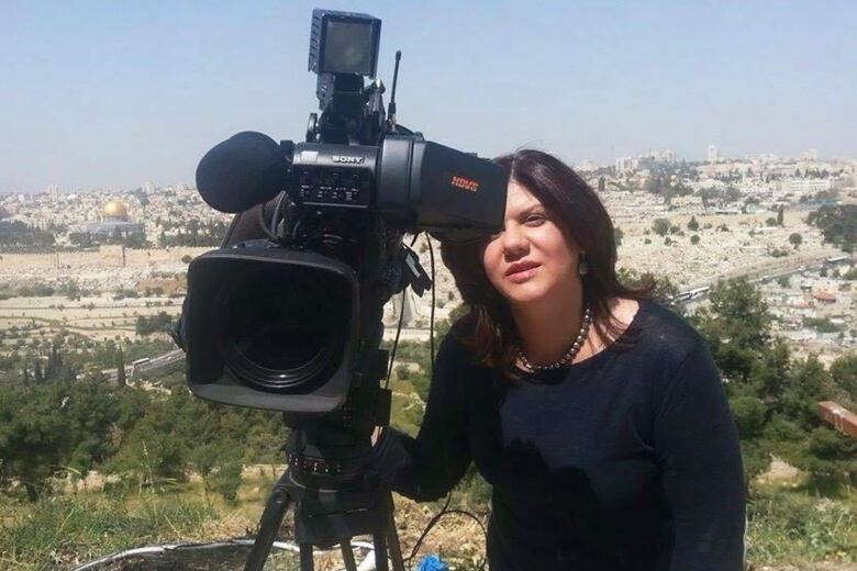 A woman wearing black looks through a TV camera in front of a background of hills and white buidlings.