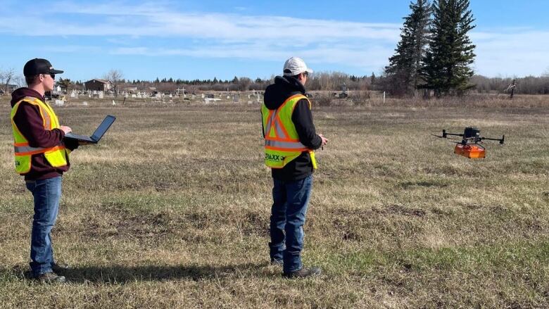 Two men in reflective vests watch as a drone hovers above a field.