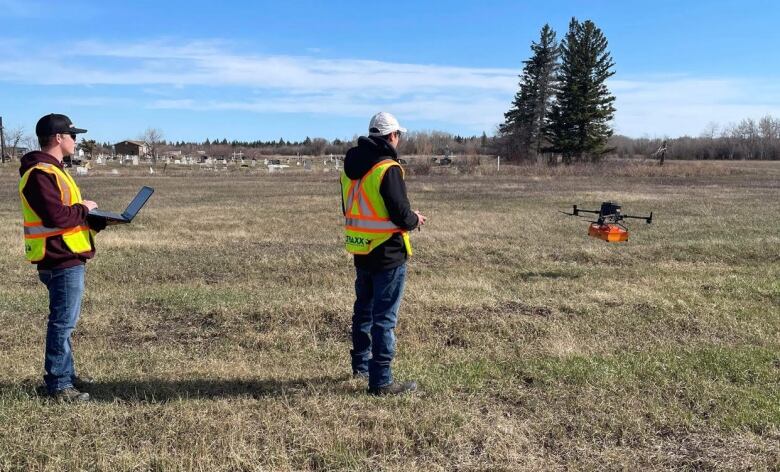 Two men in reflective vests watch as a drone hovers above a field.