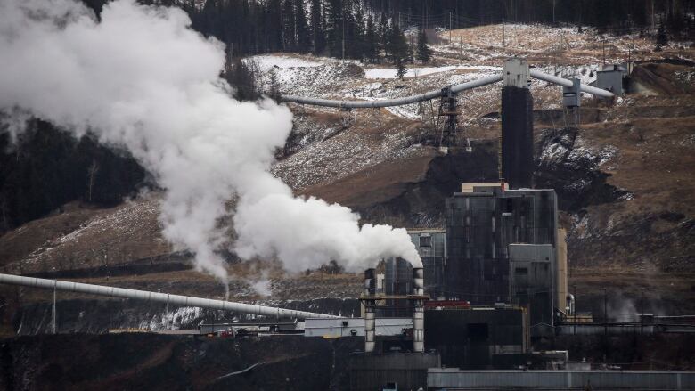 Smokestacks are emitting fumes at a coal mining facility, in the medium distance. Mountains with flecks of snow are visible in the background.