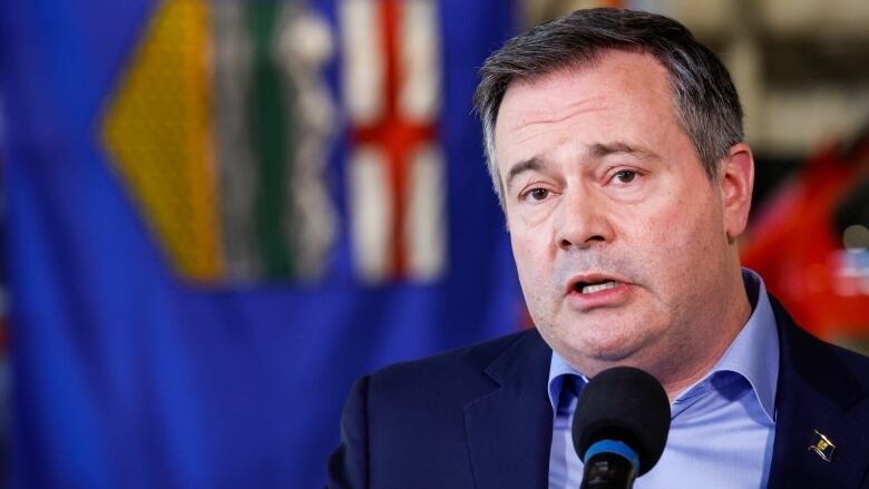 A man in a blue suit stands before the Alberta flag. 