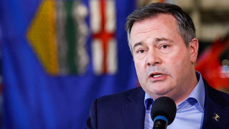 A man in a blue suit stands before the Alberta flag. 