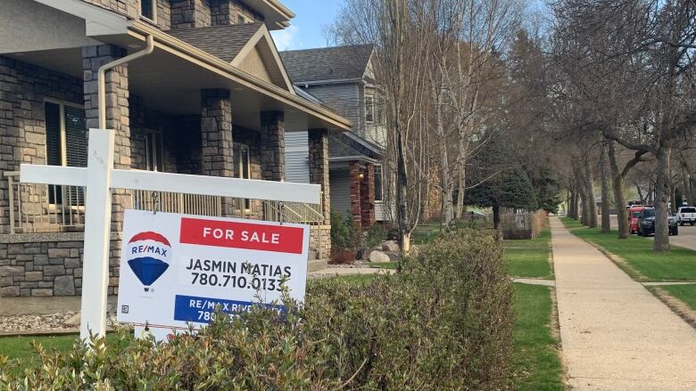 A for sale sign is seen on a home with hedges. 