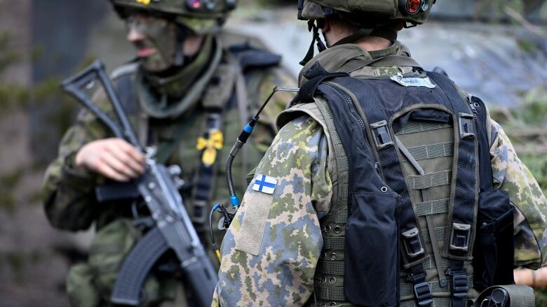 Two men in military camouflage stand, one holding an assault rifle.