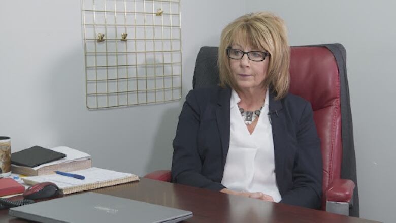 A blond woman in a grey suit with a white shirt sits behind a desk.