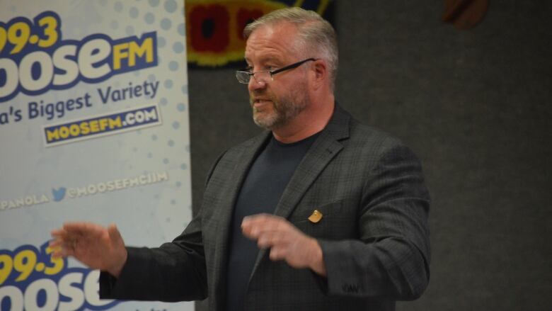 A man with grey hair and a beard wearing glasses and a blazer waves his hands while speaking to a crowd. 