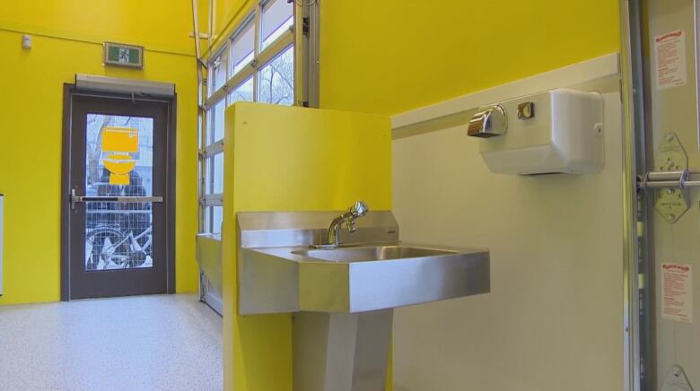 A stainless steel sink and a hand dryer are shown inside a washroom with bright yellow walls.