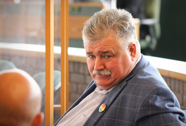 A man with short grey hair and a grey moustache wearing a blue plaid jacket and white shirt looks up.