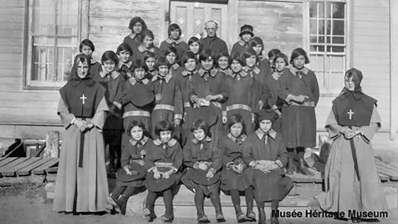 A black-and-white photograph of Indigenous children with teachers and a priest in front of a wooden building.