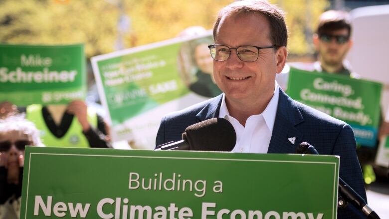 Man at podium with green and white signs behind him