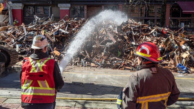 Two firefighters spray water onto the rubble where a building burned down.