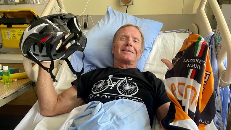 Brian Atkinson lies in a hospital bed while holding up the helmet and the jersey he was wearing when he was hit by a driver.