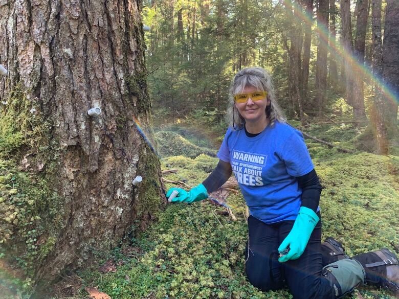 A white woman with grey hair, wearing a blue t-shirt and googles is seen next to a ;arge tree trunk.