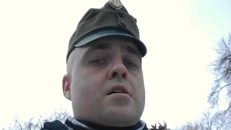 A man wearing a military-style cap and a winter jacket stands outside, looking down at the camera.