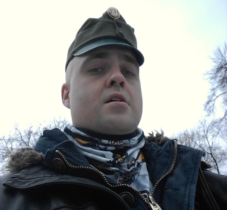 A man wearing a military-style cap and a winter jacket stands outside, looking down at the camera.