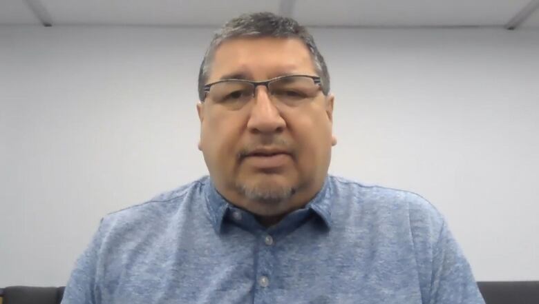 A man with grey hair wearing glasses and a blue shirt is pictured against a white wall.