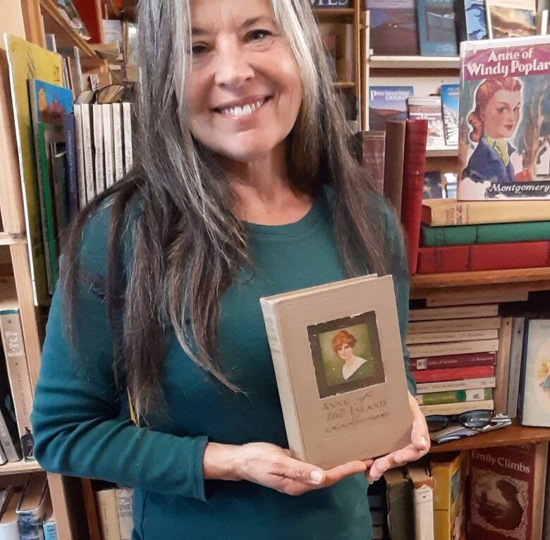 Smiling woman holds book with title Anne of the Island as she stands in front of bookshelves.