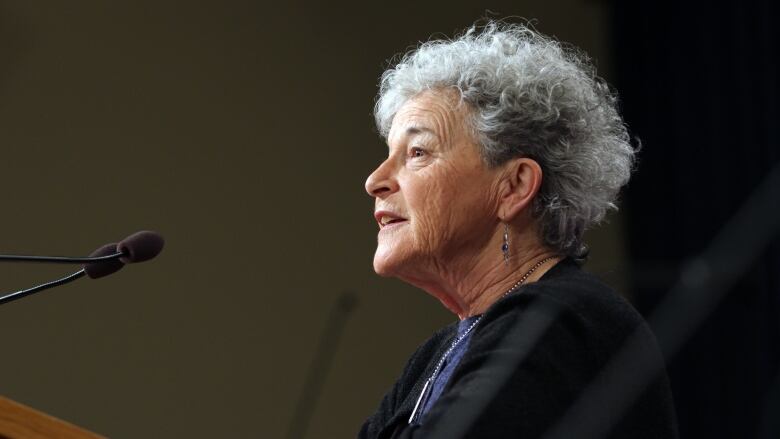 A woman with white, curly hair speaks into a mic at a podium. 