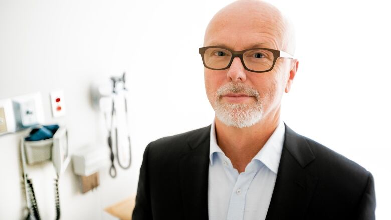 Blading man in glasses wearing a black jacket and open-neck white dress shirt, standing in a medical office.