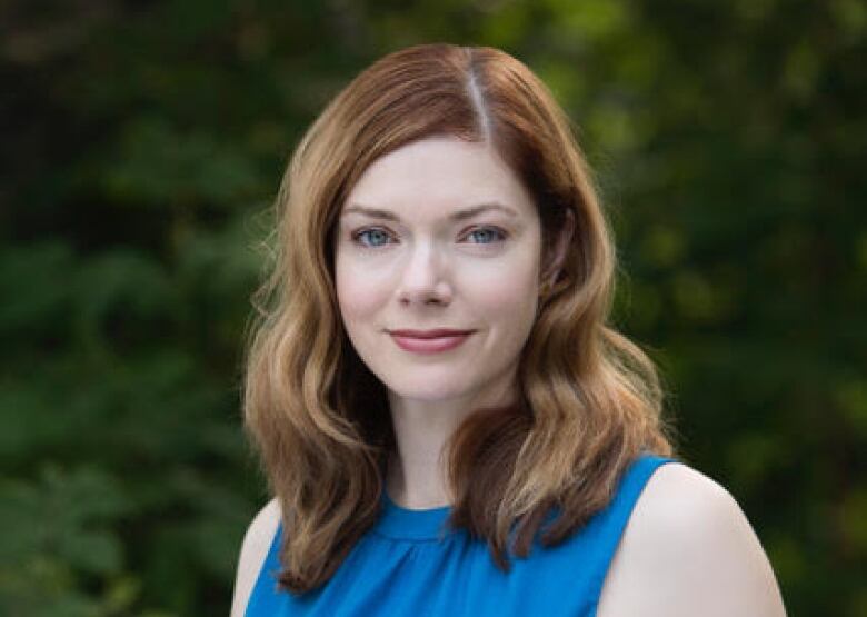 Headshot of woman with red hair, wearing a blue dress, with trees in the background.