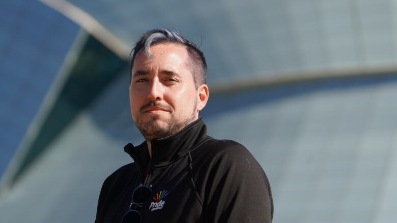 A man with short hair and a black sweater that reads 'Pride Winnipeg' stands in front of a building.