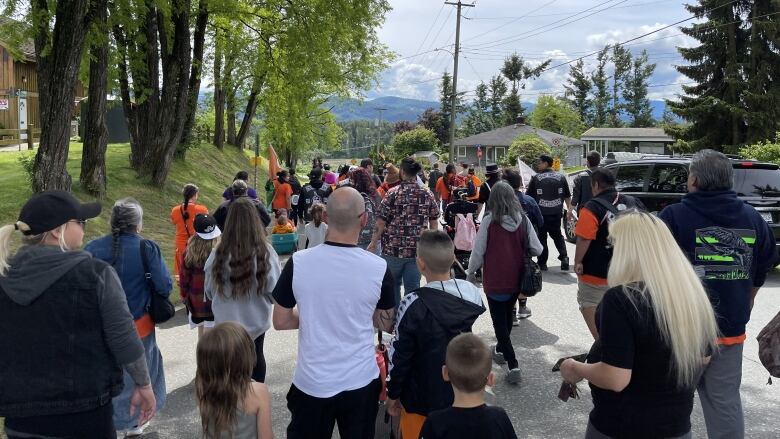 Marchers are seen on a side road. Some of them are wearing high visibility vests. All of them have their back to the camera. A child is visible holding a woman's hand.