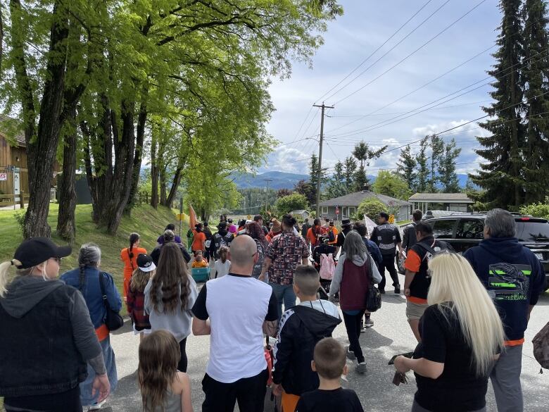 Marchers are seen on a side road. Some of them are wearing high visibility vests. All of them have their back to the camera. A child is visible holding a woman's hand.