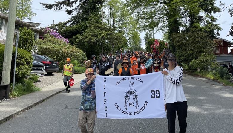 Marchers are seen coming towards the camera, with one man in a high-vis vest behind them. They are holding up a banner that reads: 