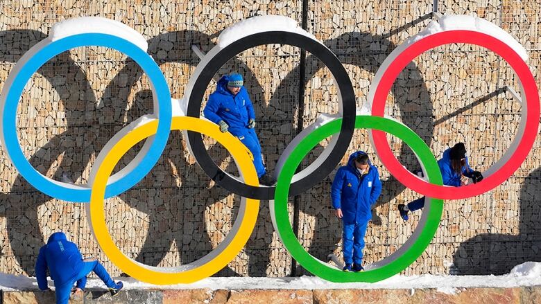 A large display of Olympic rings, partially covered in snow, is scaled by people wearing blue snowsuits.