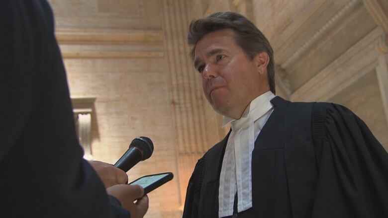 Man in black lawyer's gown talks to reporters in courthouse lobby