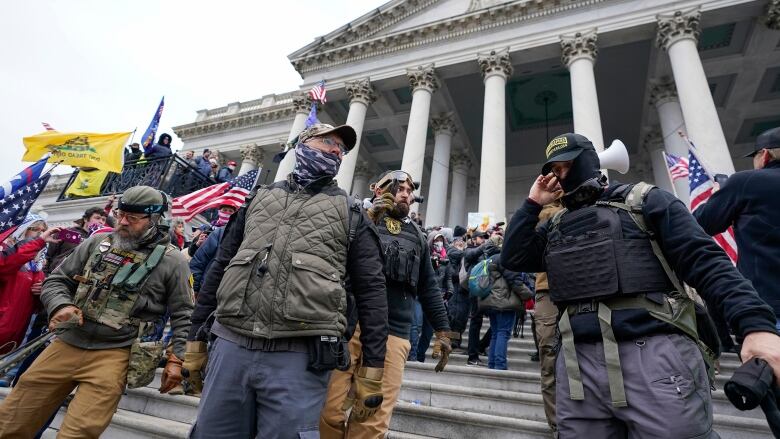 A large group of men  some masked and wearing tactical gear  stand on the steps of a government building. 