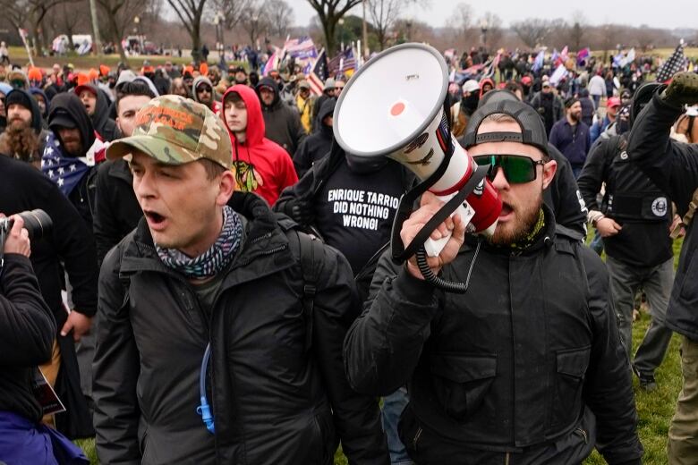 Two men in black jackets stand in front of a group of people, one with sunglasses speaks into a bull horn.