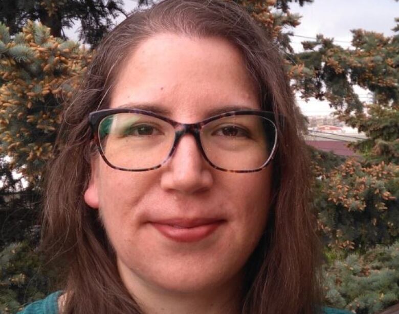 Woman with glasses standing outside in front of conifers. 