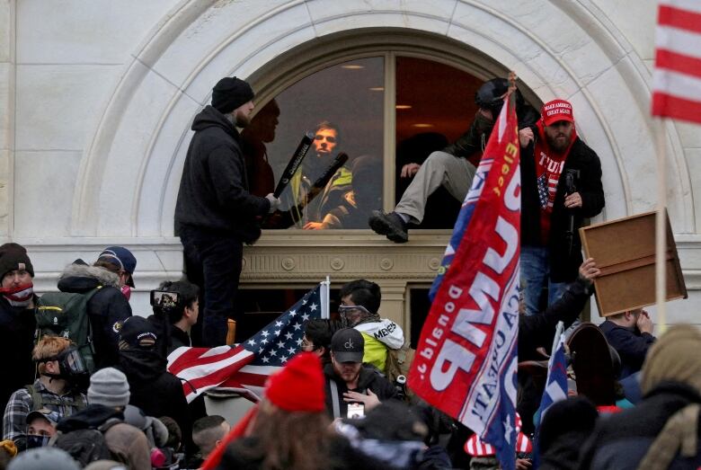 Crowds gathered in front of doorway, holding signs and U.S. flag, one man climbs through window, another holds a baseball bat.