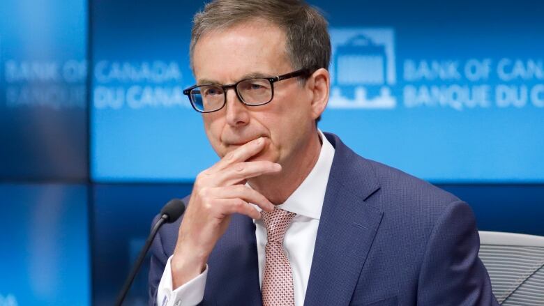 A man wearing dark-framed glasses, a blue suit and a patterned pink tie sits in front of a microphone.