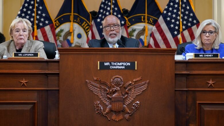 A man and two women sit behind raised desks, with American flags in the background.