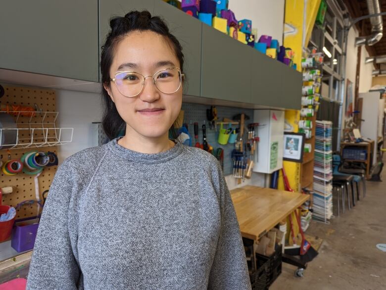 A woman in a grey sweater stands in front of an art studio wall with tools hanging up behind her.