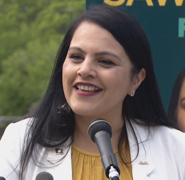 A woman with long black hair speaks into several microphones. She is standing in front of a giant photo of herself.