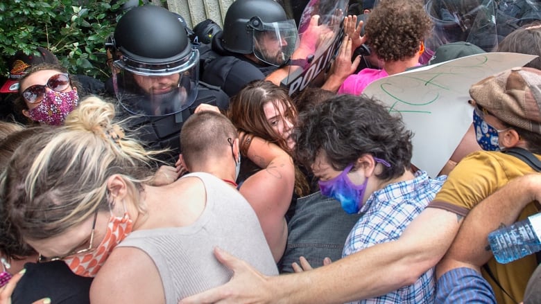 A crowd of people wrap arms around each other as they stand against police in black helmets carrying riot gear.