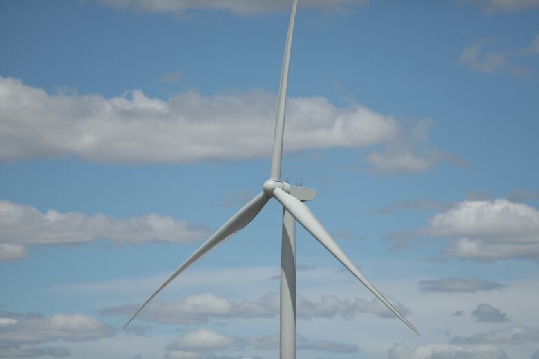 The top of a wind turbine is seen with blue sky and clouds in the background.